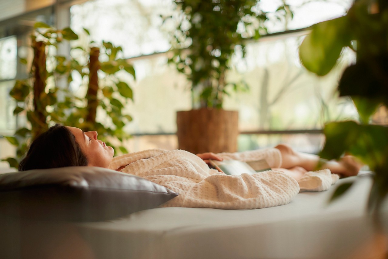 Woman reclines on a lounge chair, relaxing with eyes closed, wearing a waffle robe, in a sunlit indoor garden filled with potted green plants and large windows.